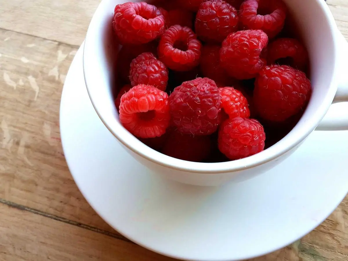 A white cup of ripe raspberries on a wooden table